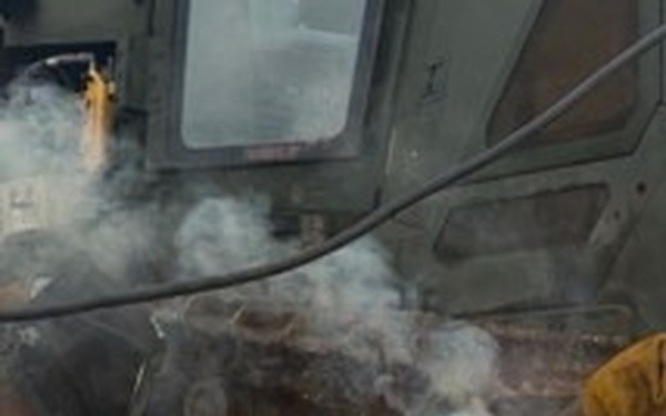 A worker is performing welding or cutting operations on a large piece of heavy metal equipment or machinery, likely outdoors or in an industrial yard. Bright arc light, colorful flame, smoke, and sparks are visible at the work point. The worker is wearing protective leather gloves and appears to be holding a metal component against the workpiece.