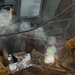 A worker is performing welding or cutting operations on a large piece of heavy metal equipment or machinery, likely outdoors or in an industrial yard. Bright arc light, colorful flame, smoke, and sparks are visible at the work point. The worker is wearing protective leather gloves and appears to be holding a metal component against the workpiece.