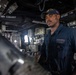 A U.S. Navy sailor in dark blue coveralls stands in what appears to be a mechanical or engineering space aboard a naval vessel. The sailor is wearing a Navy ball cap with ship insignia and a name tag reading 'Timothy Smith' with the number '76' visible on the uniform. The space contains various mechanical equipment, control panels, and labeled ventilation ducts marked 'EXHAUST' and 'INTAKE'.
