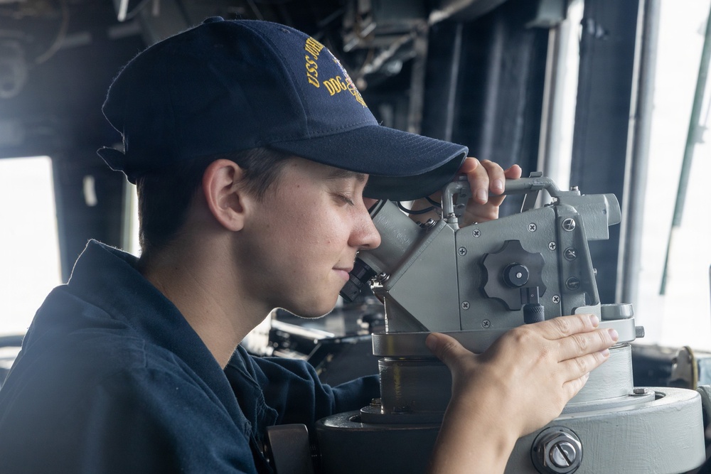 A U.S. Navy sailor is using a gyrocompass or bearing circle instrument on the bridge of a naval vessel. The sailor is wearing a navy blue uniform and a USS ship cap with 'DDG' visible in yellow lettering. The image captures a navigation or watchstanding duty being performed aboard what appears to be a destroyer-class warship.