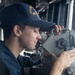 A U.S. Navy sailor is using a gyrocompass or bearing circle instrument on the bridge of a naval vessel. The sailor is wearing a navy blue uniform and a USS ship cap with 'DDG' visible in yellow lettering. The image captures a navigation or watchstanding duty being performed aboard what appears to be a destroyer-class warship.