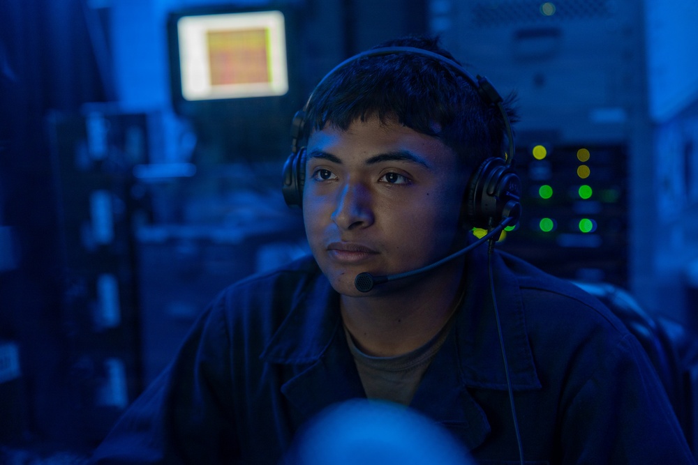 A young person wearing a military-style headset with boom microphone is seated at what appears to be a command or operations station. The environment is bathed in blue ambient lighting, characteristic of a naval combat information center or similar military operations room. Multiple illuminated screens and indicator lights are visible in the background.