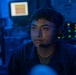 A young person wearing a military-style headset with boom microphone is seated at what appears to be a command or operations station. The environment is bathed in blue ambient lighting, characteristic of a naval combat information center or similar military operations room. Multiple illuminated screens and indicator lights are visible in the background.