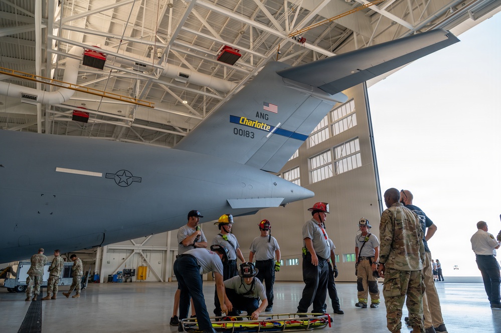 A training exercise is taking place inside a military aircraft hangar featuring a large gray military transport aircraft marked 'ANG Charlotte 00183' with a US flag insignia. Firefighters wearing helmets and military personnel in camouflage uniforms are gathered beneath the aircraft's tail section. The scene appears to be an aircraft rescue and firefighting (ARFF) or emergency response training drill.