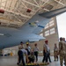 A training exercise is taking place inside a military aircraft hangar featuring a large gray military transport aircraft marked 'ANG Charlotte 00183' with a US flag insignia. Firefighters wearing helmets and military personnel in camouflage uniforms are gathered beneath the aircraft's tail section. The scene appears to be an aircraft rescue and firefighting (ARFF) or emergency response training drill.