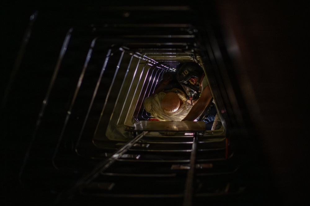 A worker wearing a respirator mask and helmet is visible inside a confined, tunnel-like metal shaft or duct, viewed from below looking upward. The structure appears to be a rectangular metal conduit with ribbed or corrugated walls, possibly a ventilation shaft, tank interior, or industrial access tunnel. The worker appears to be performing inspection or maintenance work in a confined space.