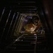 A worker wearing a respirator mask and helmet is visible inside a confined, tunnel-like metal shaft or duct, viewed from below looking upward. The structure appears to be a rectangular metal conduit with ribbed or corrugated walls, possibly a ventilation shaft, tank interior, or industrial access tunnel. The worker appears to be performing inspection or maintenance work in a confined space.