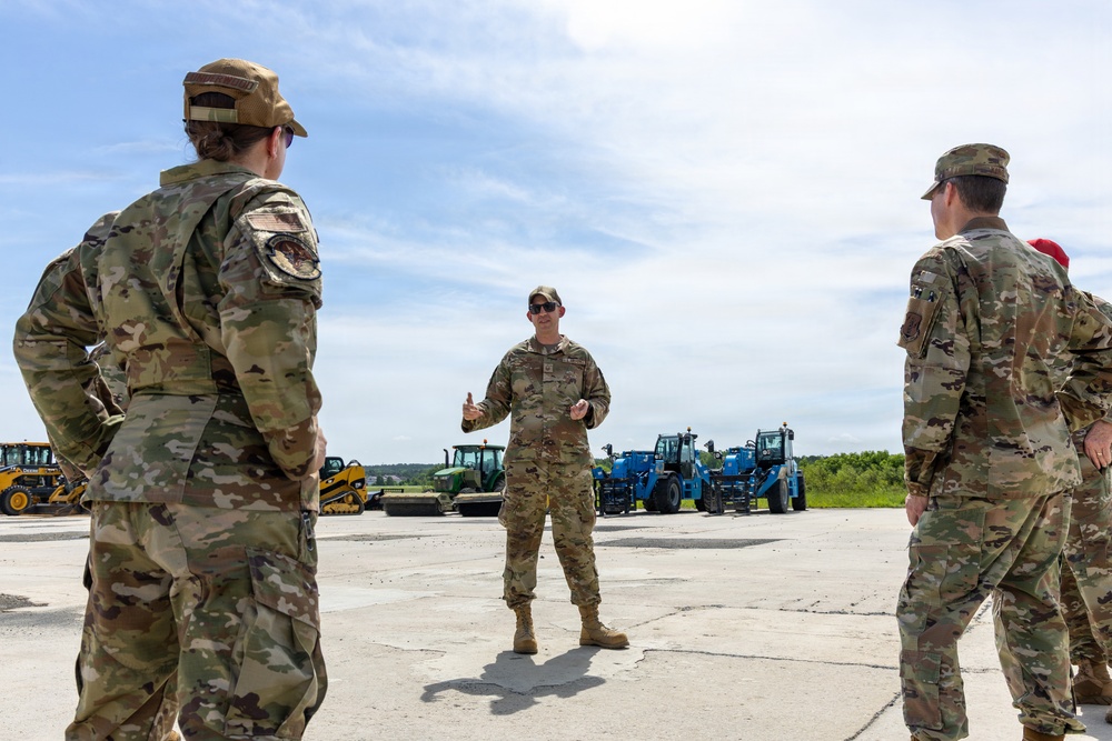 A group of military personnel in OCP (Operational Camouflage Pattern) uniforms are gathered on a concrete surface, likely a flight line or airfield apron. One individual in the center appears to be briefing or instructing the others, gesturing with their hands. Various heavy construction and ground support equipment vehicles are visible in the background.