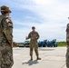 A group of military personnel in OCP (Operational Camouflage Pattern) uniforms are gathered on a concrete surface, likely a flight line or airfield apron. One individual in the center appears to be briefing or instructing the others, gesturing with their hands. Various heavy construction and ground support equipment vehicles are visible in the background.