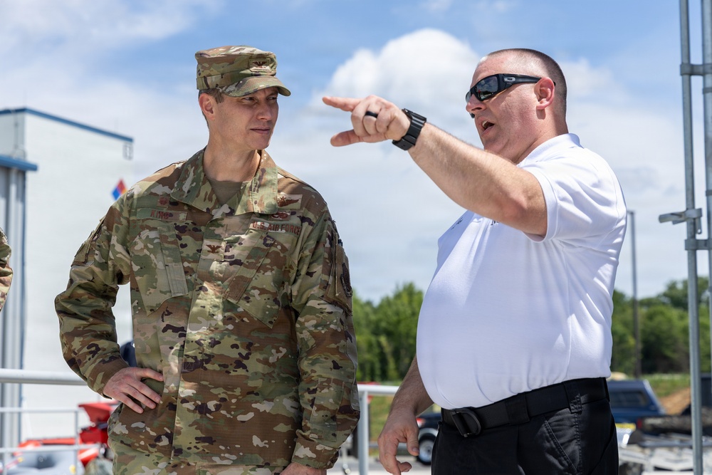 A U.S. Air Force officer in OCP (Operational Camouflage Pattern) uniform is engaged in a conversation with a civilian or plainclothes individual wearing a white polo shirt. The civilian is pointing in a specific direction, apparently briefing or directing the military officer's attention toward something off-frame. The setting appears to be an outdoor facility or installation on a clear, partly cloudy day.