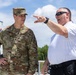 A U.S. Air Force officer in OCP (Operational Camouflage Pattern) uniform is engaged in a conversation with a civilian or plainclothes individual wearing a white polo shirt. The civilian is pointing in a specific direction, apparently briefing or directing the military officer's attention toward something off-frame. The setting appears to be an outdoor facility or installation on a clear, partly cloudy day.