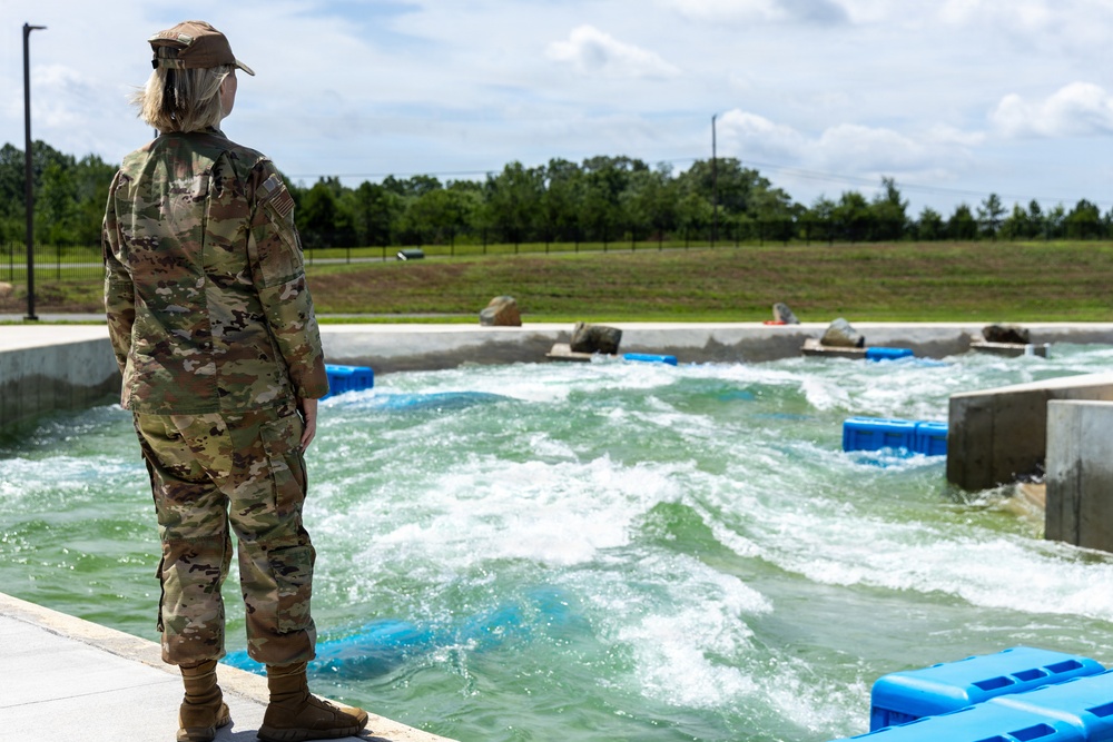 A female soldier in US Army OCP (Operational Camouflage Pattern) uniform stands at the edge of what appears to be a whitewater training or water obstacle course facility. She is observing the churning water from a concrete platform, with blue flotation barriers and concrete structures visible in the water. The setting appears to be a military training installation with green fields and trees in the background.