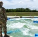 A female soldier in US Army OCP (Operational Camouflage Pattern) uniform stands at the edge of what appears to be a whitewater training or water obstacle course facility. She is observing the churning water from a concrete platform, with blue flotation barriers and concrete structures visible in the water. The setting appears to be a military training installation with green fields and trees in the background.