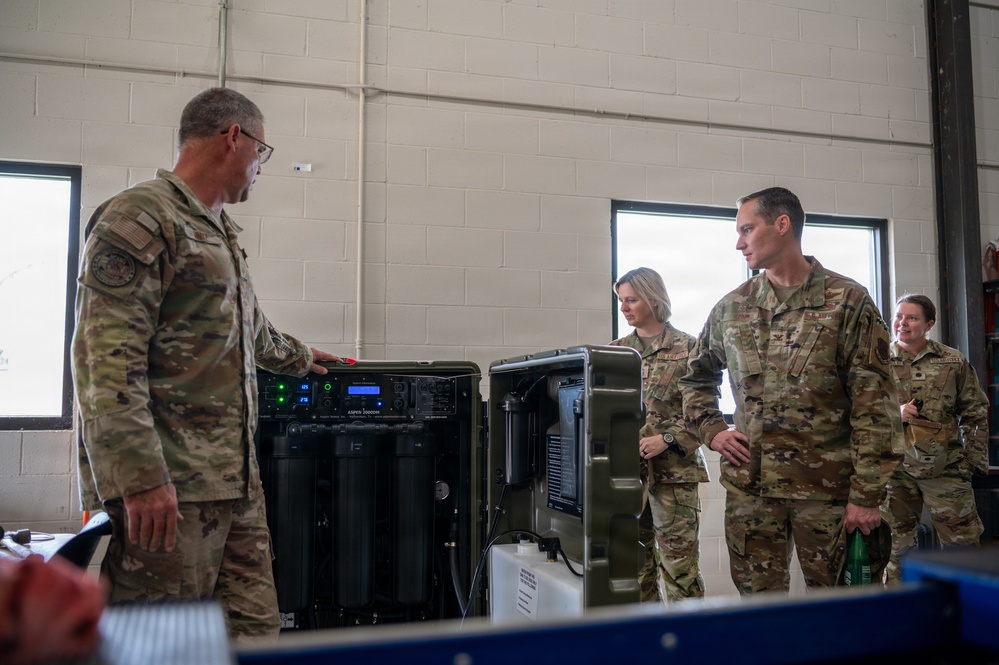 Military personnel in OCP (Operational Camouflage Pattern) uniforms are gathered around what appears to be a water purification or filtration system inside a facility with cinder block walls. One individual on the left appears to be briefing or demonstrating the equipment to the group. The setting appears to be a maintenance bay or equipment storage facility on a military installation.