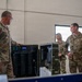 Military personnel in OCP (Operational Camouflage Pattern) uniforms are gathered around what appears to be a water purification or filtration system inside a facility with cinder block walls. One individual on the left appears to be briefing or demonstrating the equipment to the group. The setting appears to be a maintenance bay or equipment storage facility on a military installation.