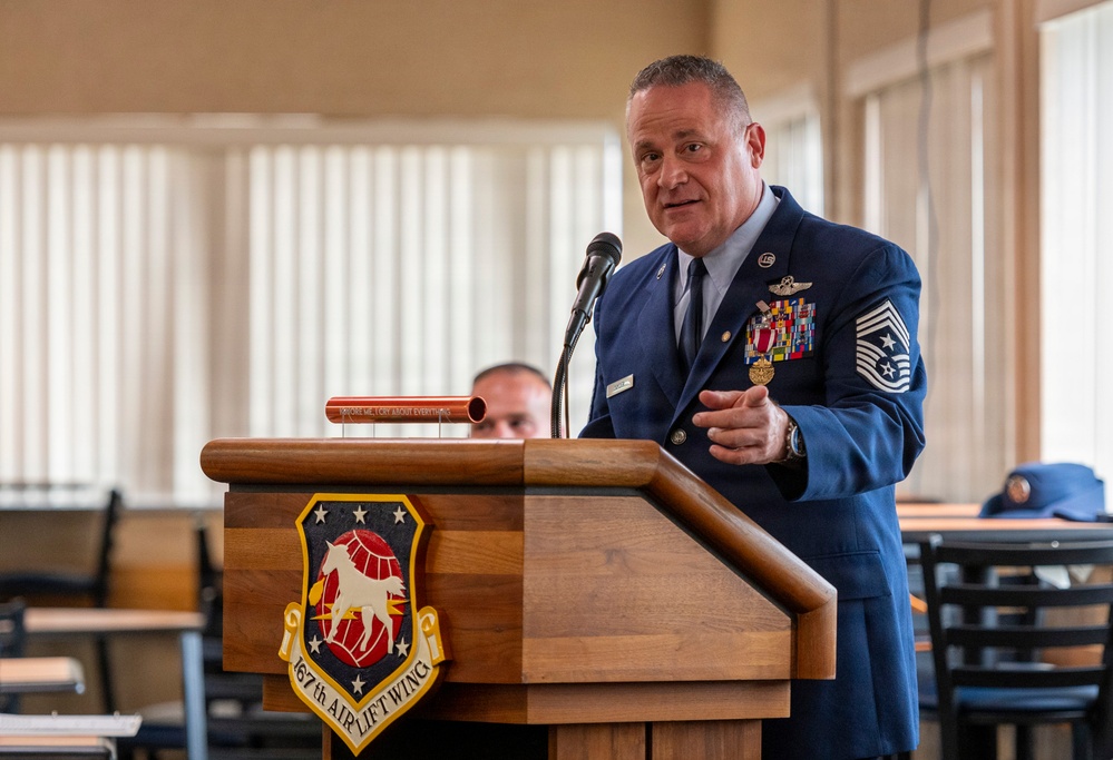A senior U.S. Air Force enlisted member in dress blues is delivering a speech at a wooden podium bearing the insignia of the 467th Airlift Wing. The individual displays numerous service ribbons, medals, and what appears to be a Command Chief Master Sergeant rank insignia. The setting appears to be a formal military ceremony held in a dining facility or meeting hall.