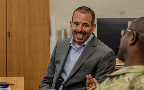 A civilian in a gray blazer and light blue shirt is engaged in a conversation with a U.S. Army soldier in MultiCam (OCP) uniform. The setting appears to be a military office or administrative workspace. The civilian is smiling broadly, suggesting a positive and engaged interaction.
