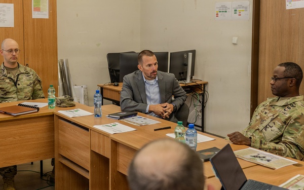 A briefing or meeting is taking place in what appears to be a military facility, with several U.S. Army personnel in camouflage uniforms and at least two civilians seated around wooden desks arranged in a U-shape. A civilian in a gray blazer appears to be leading or presenting to the group. A U.S. flag is visible in the background, suggesting an official military setting.