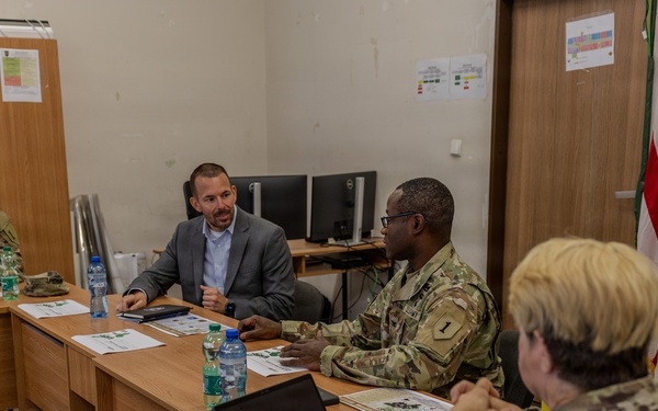 A briefing or meeting is taking place in a military facility between uniformed U.S. Army personnel and at least one civilian in a gray suit. Participants are seated around wooden tables arranged in a U-shape, with documents, water bottles, and a tablet visible on the surfaces. A U.S. flag is displayed in the background near a door.