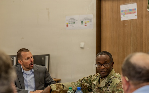 A meeting is taking place in a conference room setting with a U.S. Army soldier in camouflage uniform and a civilian in a gray suit engaged in discussion across a wooden table. Several other attendees are visible from behind in the foreground. An American flag is visible in the background along with posted documents on the wall.