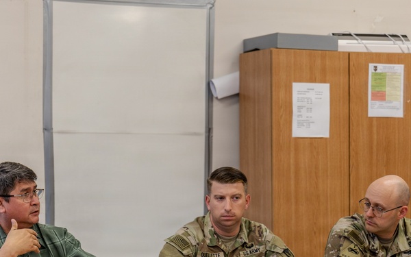 Three individuals are seated at a wooden table in what appears to be a briefing or meeting room. Two of the individuals are wearing U.S. Army camouflage uniforms, while the third, on the left, is a civilian in a green plaid shirt who appears to be speaking and gesturing. The setting suggests a formal military meeting or coordination session.