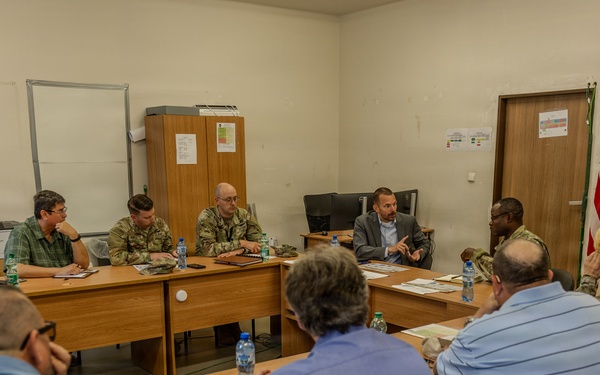 A formal meeting or briefing is taking place in what appears to be a military facility or office, with a mixed group of military personnel in camouflage uniforms and civilians in business casual attire seated around conference tables arranged in a U-shape. An American flag and what appears to be a military unit guidon or organizational flag are visible in the background. One individual in civilian clothing appears to be actively speaking or presenting to the group.
