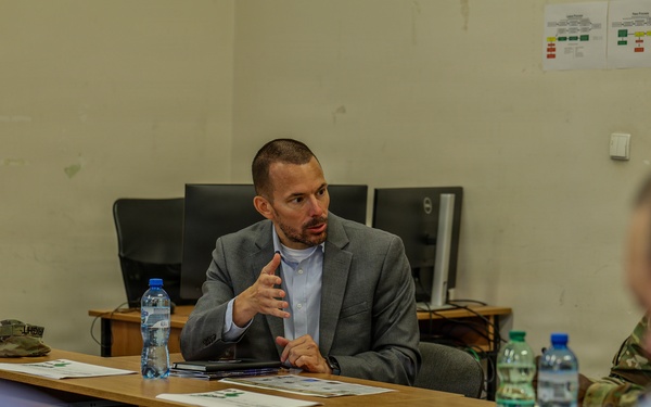 A man in a gray blazer and light blue shirt is seated at a conference table, actively speaking and gesturing with his hands during what appears to be a formal meeting or briefing. Several other individuals are seated across from him, viewed from behind, with documents and water bottles spread across the table. The setting appears to be an office or military briefing room with computers and posted charts visible in the background.