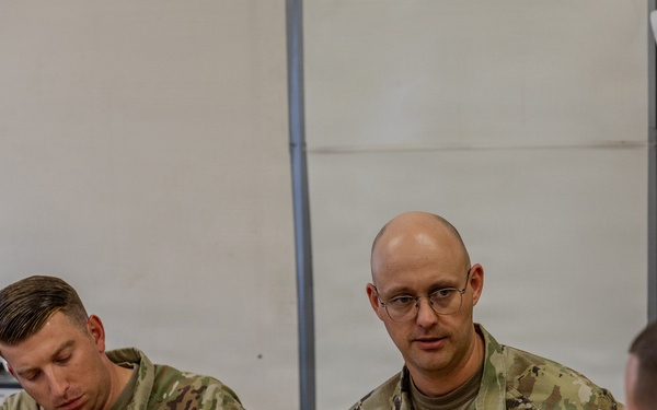 Two U.S. Army soldiers in MultiCam/OCP camouflage uniforms are seated at a table during what appears to be a meeting or briefing. The soldier on the right, identified by name tape as 'WRIGHT,' is speaking while the soldier on the left, identified as 'OUELLETTE,' is taking notes. A civilian in a suit jacket is seated across the table, suggesting an official or diplomatic meeting context.