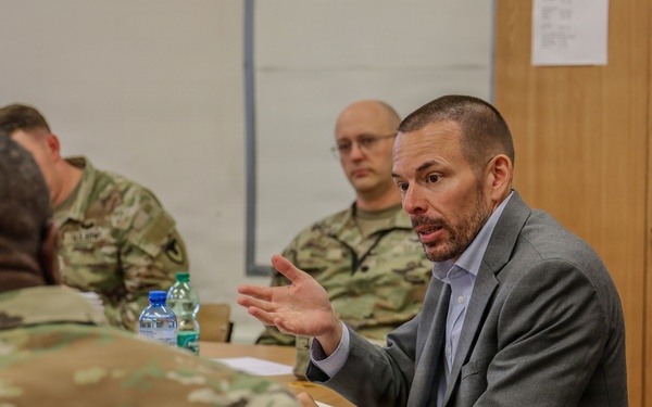 A civilian in a gray suit is engaged in an active discussion with military personnel wearing camouflage uniforms, likely U.S. Army OCP (Operational Camouflage Pattern) uniforms. The setting appears to be a briefing room or conference room, suggesting a formal meeting or training session. The civilian appears to be presenting or explaining something, as indicated by his hand gestures.