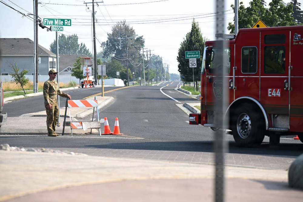 A road closure or checkpoint scene at the intersection of Five Mile Road in Spokane, Washington. A person in military/camouflage clothing stands next to orange and white barricades with traffic cones, while a red fire engine labeled 'E44' is positioned on the right side of the frame. The road ahead appears empty and newly paved, with a 'Welcome to Spokane' speed limit 30 sign visible in the distance.