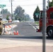 A road closure or checkpoint scene at the intersection of Five Mile Road in Spokane, Washington. A person in military/camouflage clothing stands next to orange and white barricades with traffic cones, while a red fire engine labeled 'E44' is positioned on the right side of the frame. The road ahead appears empty and newly paved, with a 'Welcome to Spokane' speed limit 30 sign visible in the distance.