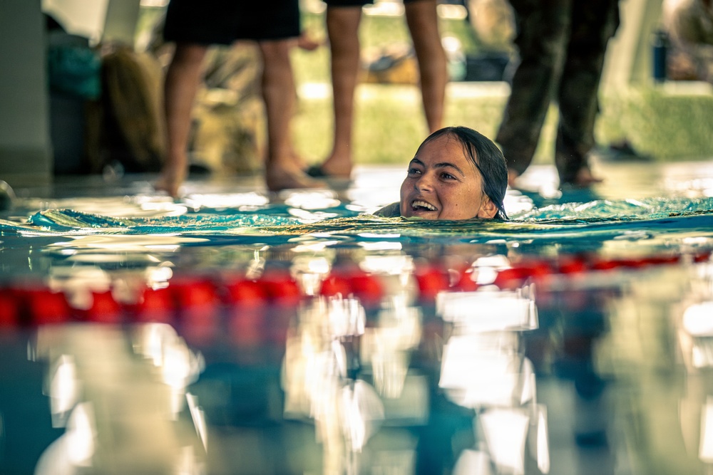 A smiling woman is swimming in a pool, her head just above the water surface near a lane divider with red and white floats. Several people are standing at the pool's edge in the background, visible only from the waist down. The image is taken from a low, water-level perspective, creating a dramatic and intimate composition.