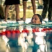 A smiling woman is swimming in a pool, her head just above the water surface near a lane divider with red and white floats. Several people are standing at the pool's edge in the background, visible only from the waist down. The image is taken from a low, water-level perspective, creating a dramatic and intimate composition.