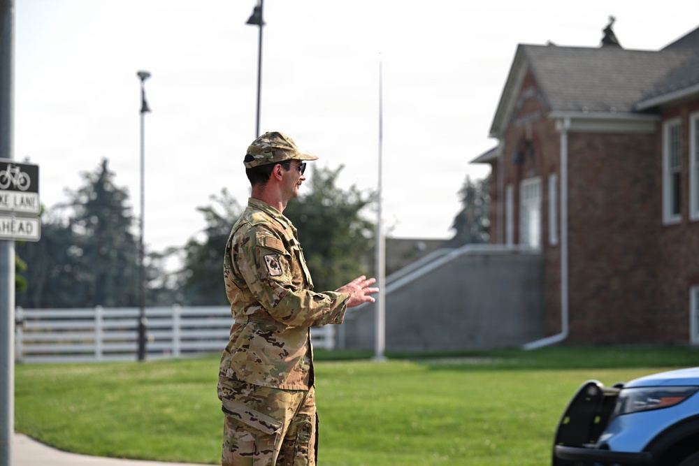 A military service member in OCP (Operational Camouflage Pattern) uniform is standing outdoors on what appears to be a military installation, gesturing with their hands as if speaking or directing. The background includes a brick building, a white fence, a flagpole, and a partial view of a law enforcement or security vehicle. The setting suggests an administrative or garrison area of a military base.