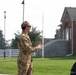 A military service member in OCP (Operational Camouflage Pattern) uniform is standing outdoors on what appears to be a military installation, gesturing with their hands as if speaking or directing. The background includes a brick building, a white fence, a flagpole, and a partial view of a law enforcement or security vehicle. The setting suggests an administrative or garrison area of a military base.