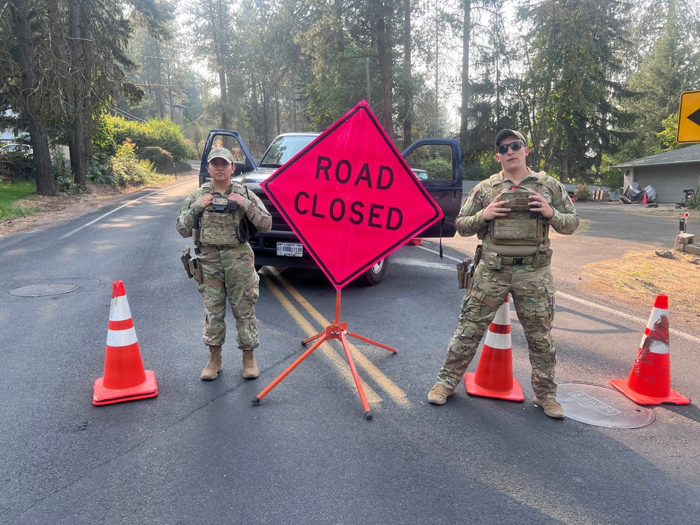 Two military or law enforcement personnel in camouflage uniforms and tactical vests are standing at a road closure checkpoint. They are flanking a bright orange diamond-shaped 'ROAD CLOSED' sign mounted on an orange stand, surrounded by traffic cones. The setting appears to be a residential or rural road lined with tall conifer trees, with hazy air suggesting possible wildfire smoke in the area.
