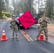 Two military or law enforcement personnel in camouflage uniforms and tactical vests are standing at a road closure checkpoint. They are flanking a bright orange diamond-shaped 'ROAD CLOSED' sign mounted on an orange stand, surrounded by traffic cones. The setting appears to be a residential or rural road lined with tall conifer trees, with hazy air suggesting possible wildfire smoke in the area.