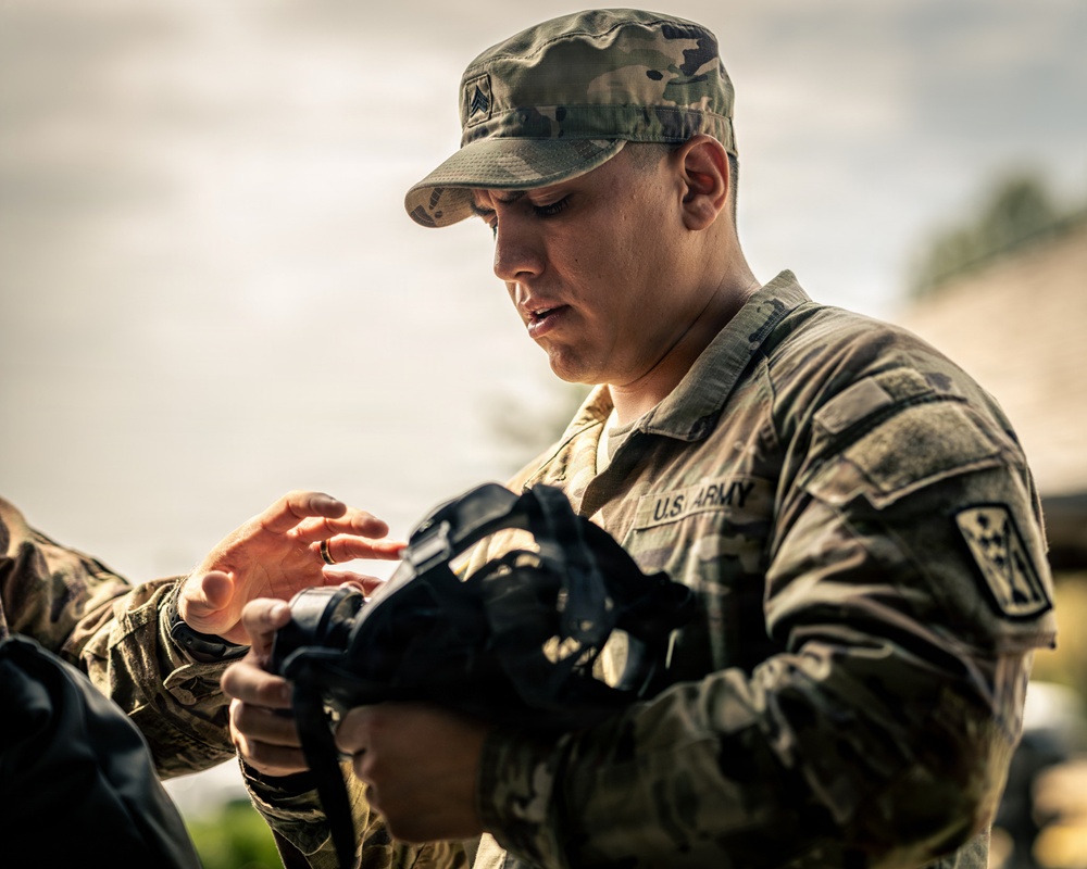 A U.S. Army soldier in Operational Camouflage Pattern (OCP) uniform is assisting another soldier with equipment, appearing to adjust or inspect a piece of gear. The soldier wears a rank insignia on their patrol cap and a unit patch on the right sleeve. The scene appears to be an outdoor training or preparation environment.