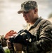 A U.S. Army soldier in Operational Camouflage Pattern (OCP) uniform is assisting another soldier with equipment, appearing to adjust or inspect a piece of gear. The soldier wears a rank insignia on their patrol cap and a unit patch on the right sleeve. The scene appears to be an outdoor training or preparation environment.