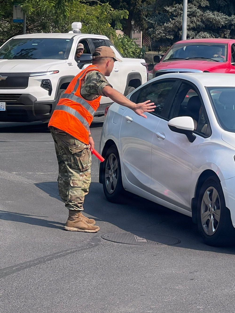 A person wearing military camouflage uniform and an orange high-visibility safety vest is directing traffic at what appears to be a busy intersection or checkpoint. They are holding a red traffic baton in one hand while extending the other hand toward a silver sedan, signaling it to stop or slow down. Multiple vehicles including a white pickup truck and a red vehicle are visible in the background.