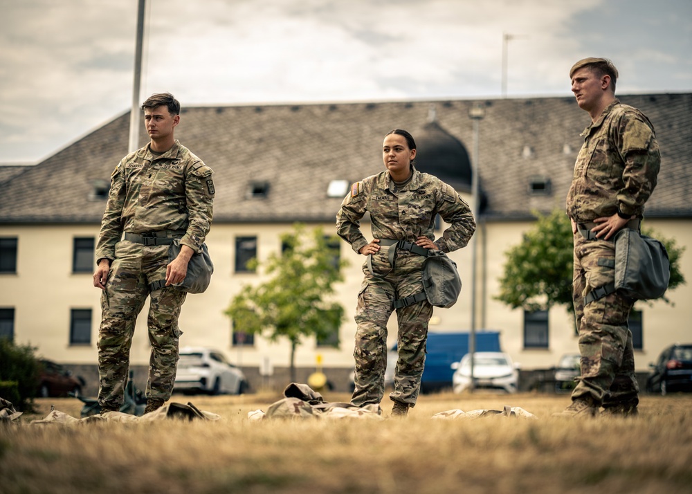 Three U.S. Army soldiers in OCP (Operational Camouflage Pattern) uniforms stand at attention on a dry grass field, with military equipment bags visible on the ground near their feet. The soldiers appear to be at a military installation, likely in Europe based on the architectural style of the building in the background. Two male soldiers flank a female soldier in the center, all appearing to be awaiting instruction or observing an activity.