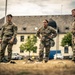 Three U.S. Army soldiers in OCP (Operational Camouflage Pattern) uniforms stand at attention on a dry grass field, with military equipment bags visible on the ground near their feet. The soldiers appear to be at a military installation, likely in Europe based on the architectural style of the building in the background. Two male soldiers flank a female soldier in the center, all appearing to be awaiting instruction or observing an activity.