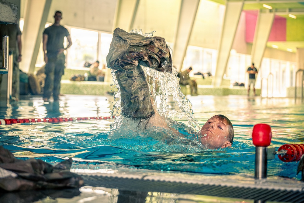 A military service member is in an indoor swimming pool performing a combat water survival training exercise, lifting waterlogged camouflage uniform clothing above the water. Several other uniformed personnel are visible in the background observing or waiting their turn. The setting appears to be a modern indoor aquatic facility with large windows and colorful wall panels.
