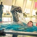 A military service member is in an indoor swimming pool performing a combat water survival training exercise, lifting waterlogged camouflage uniform clothing above the water. Several other uniformed personnel are visible in the background observing or waiting their turn. The setting appears to be a modern indoor aquatic facility with large windows and colorful wall panels.