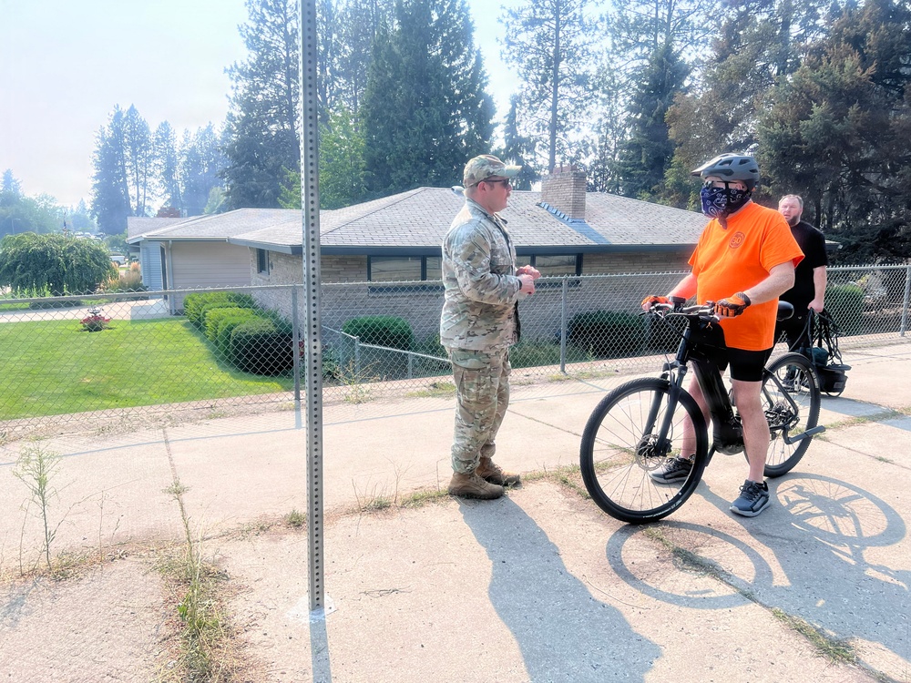 A military personnel in camouflage uniform is conversing with a civilian wearing an orange shirt, helmet, and face mask who is holding a bicycle. A third person in a black shirt is visible in the background. The scene takes place on a paved area near a residential property, with hazy/smoky skies suggesting possible nearby wildfire activity.