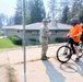 A military personnel in camouflage uniform is conversing with a civilian wearing an orange shirt, helmet, and face mask who is holding a bicycle. A third person in a black shirt is visible in the background. The scene takes place on a paved area near a residential property, with hazy/smoky skies suggesting possible nearby wildfire activity.