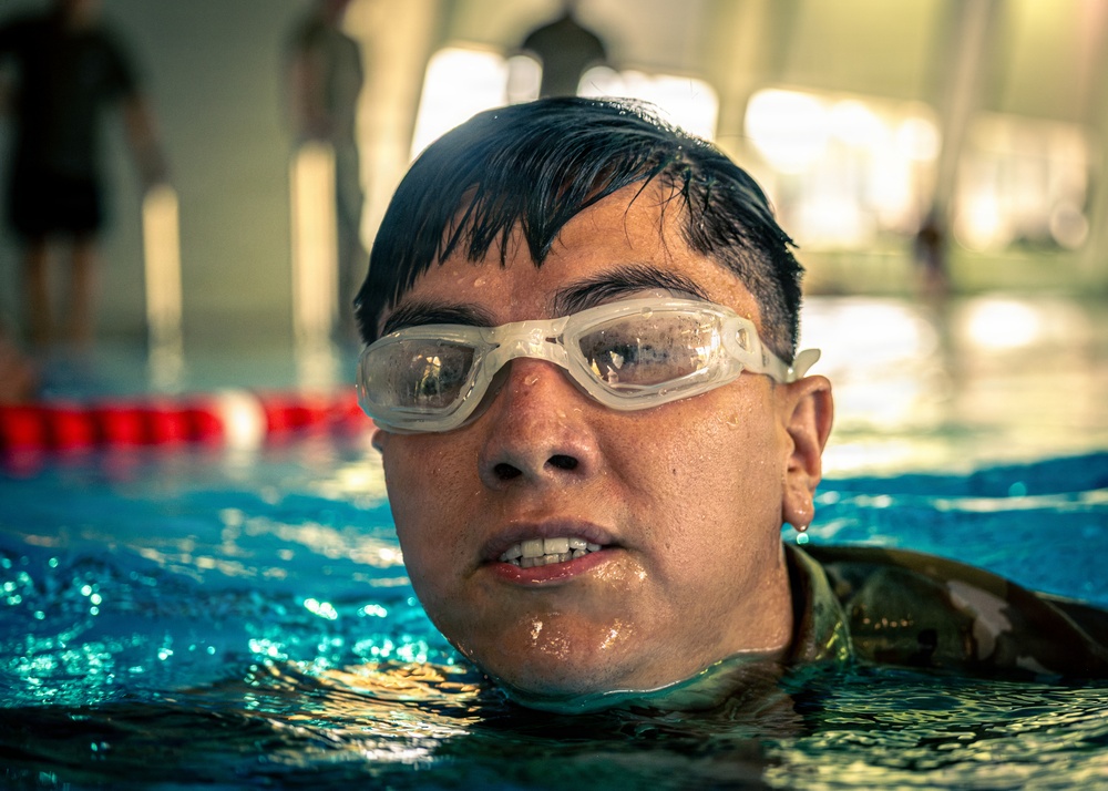 A close-up photograph of a swimmer emerging from an indoor pool, wearing clear swimming goggles pushed up on their forehead. The subject has wet dark hair and appears to be wearing a camouflage-patterned garment, suggesting possible military training. The background shows a lane-divided pool with red and white lane markers.