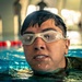 A close-up photograph of a swimmer emerging from an indoor pool, wearing clear swimming goggles pushed up on their forehead. The subject has wet dark hair and appears to be wearing a camouflage-patterned garment, suggesting possible military training. The background shows a lane-divided pool with red and white lane markers.
