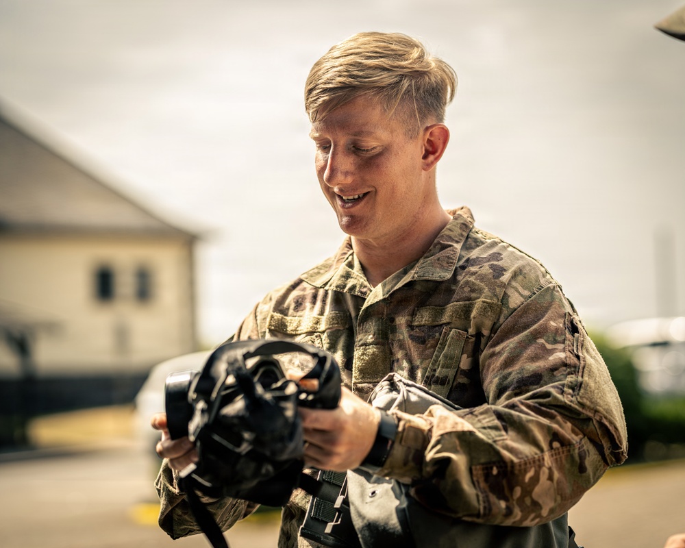 A young military service member in OCP (Operational Camouflage Pattern) uniform is examining what appears to be an M50 or similar military gas mask/protective mask outdoors. The individual appears to be smiling or speaking while inspecting the equipment. The setting appears to be a military installation or base, with buildings visible in the blurred background.