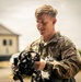 A young military service member in OCP (Operational Camouflage Pattern) uniform is examining what appears to be an M50 or similar military gas mask/protective mask outdoors. The individual appears to be smiling or speaking while inspecting the equipment. The setting appears to be a military installation or base, with buildings visible in the blurred background.